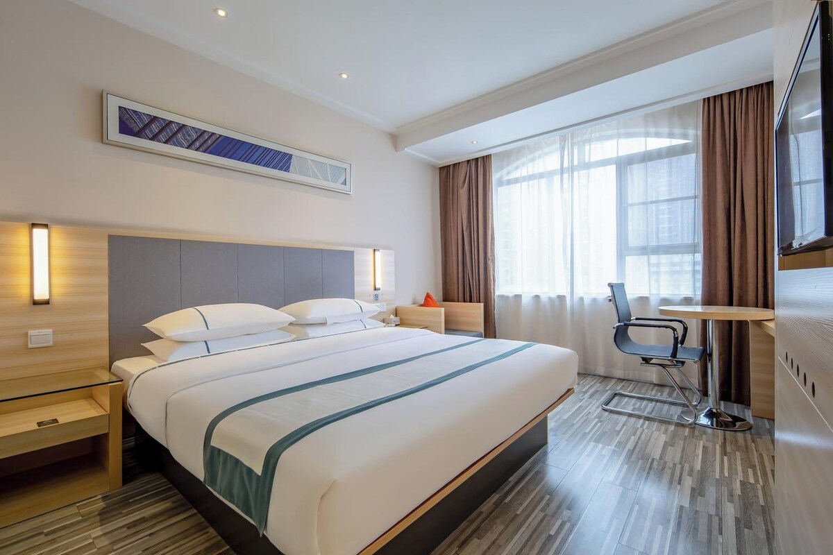 A hotel room with a large bed, bedside tables, a desk with a chair, a window with curtains, and a wall-mounted TV.