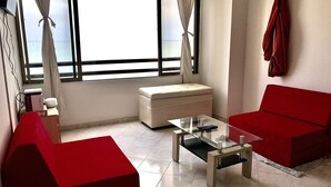 Un soggiorno con un divano rosso, un tavolino da caffè in vetro, un pouf bianco e vista mare attraverso ampie finestre.