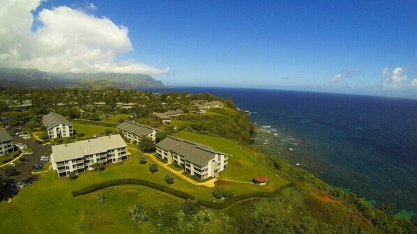 An aerial view of a coastal residential area with multiple buildings, green lawns, and a view of the ocean. - Amazing 1BR Condo in The Cliffs (Princeville)