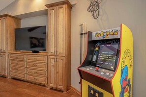 Une pièce équipée d’une borne d’arcade Pac-Man, d’un téléviseur à écran plat, de meubles en bois et d’un élément décoratif fixé au mur.