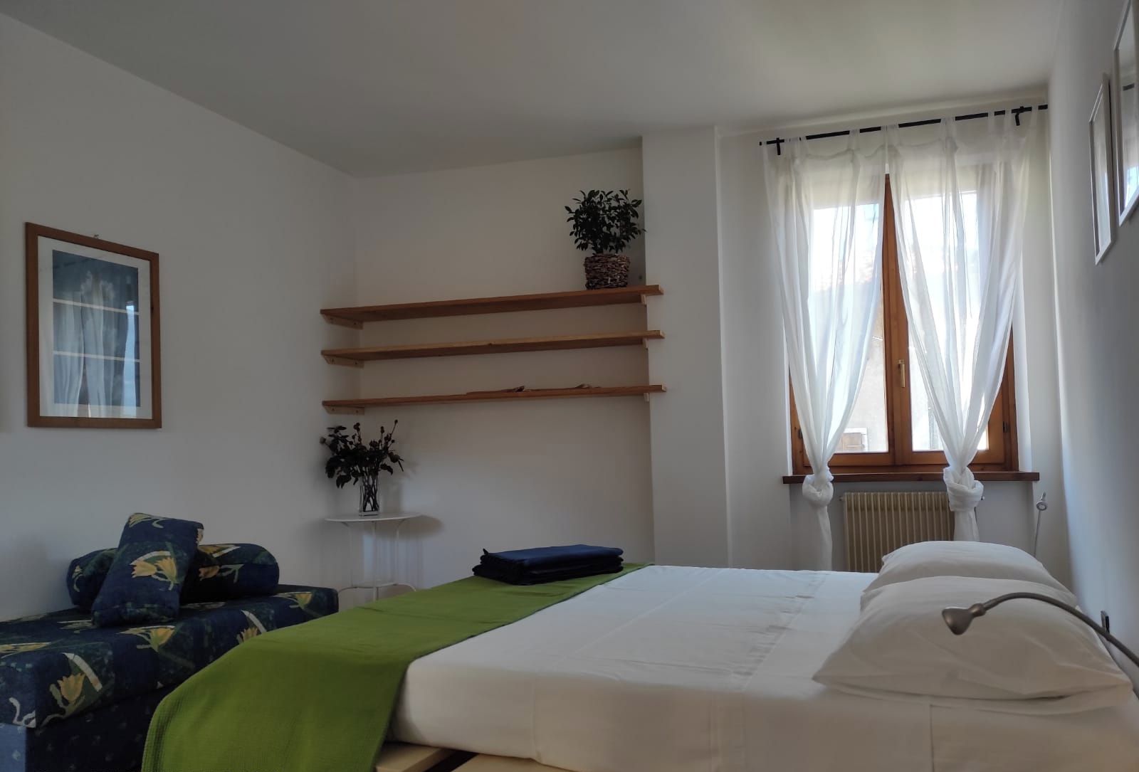Un dormitorio con cama, sofá, una mesita con planta y ventana con cortinas.