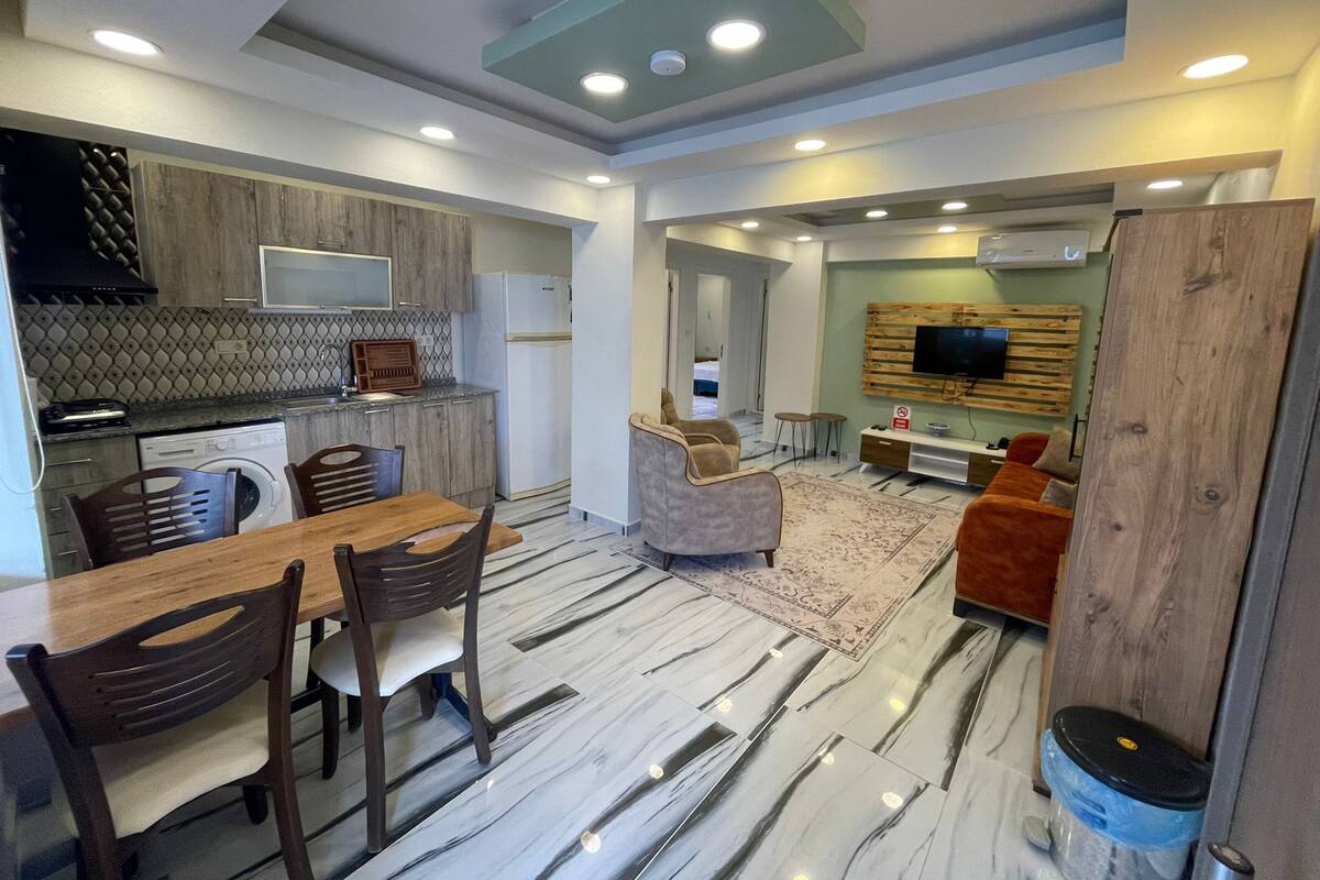 Eine moderne Küche mit integriertem Mikrowellenherd, einem Essbereich mit Holztisch und -stühlen sowie einem Wohnbereich mit Sofa und an einer Holzplatte montiertem Fernseher.