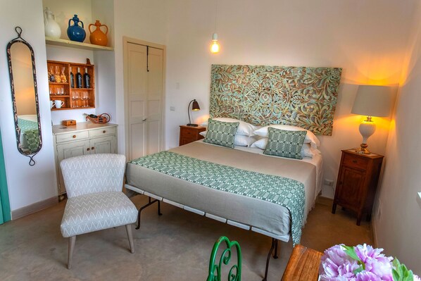 A bedroom with a bed, a chair, a nightstand, a lamp, a mirror, shelves with bottles and glasses, and a door. - Masseria Ferarelli - "Rizzeddrha" Room with Patio (Diso)