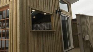 Edifici de fusta amb una finestra gran i una porta, que inclou una terrassa de fusta i una petita zona exterior.