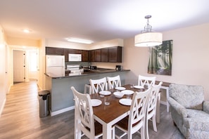 A modern kitchen with dark cabinets, a white refrigerator, and a microwave. There is a dining table with white chairs and a patterned sofa.