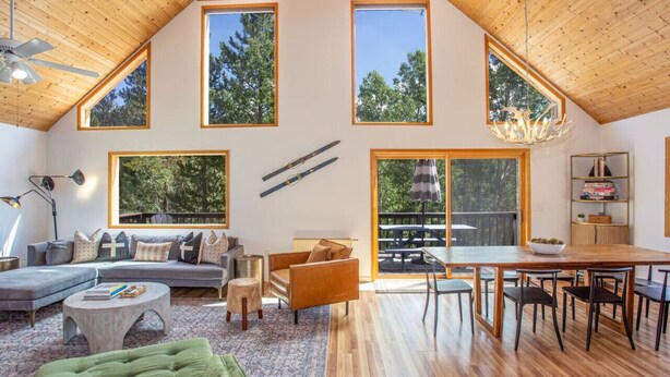 A spacious loft with high ceilings, large windows, a cozy seating area, a dining table, and a kitchen area. - Summer Cabin Retreat | Hot Tub, Firepit, Sleeps 12 (Alma)