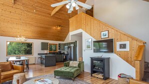 A spacious living area with a high ceiling, wooden beams, a dining table, a green sofa, and a fireplace. - Summer Cabin Retreat | Hot Tub, Firepit, Sleeps 12 (Alma)