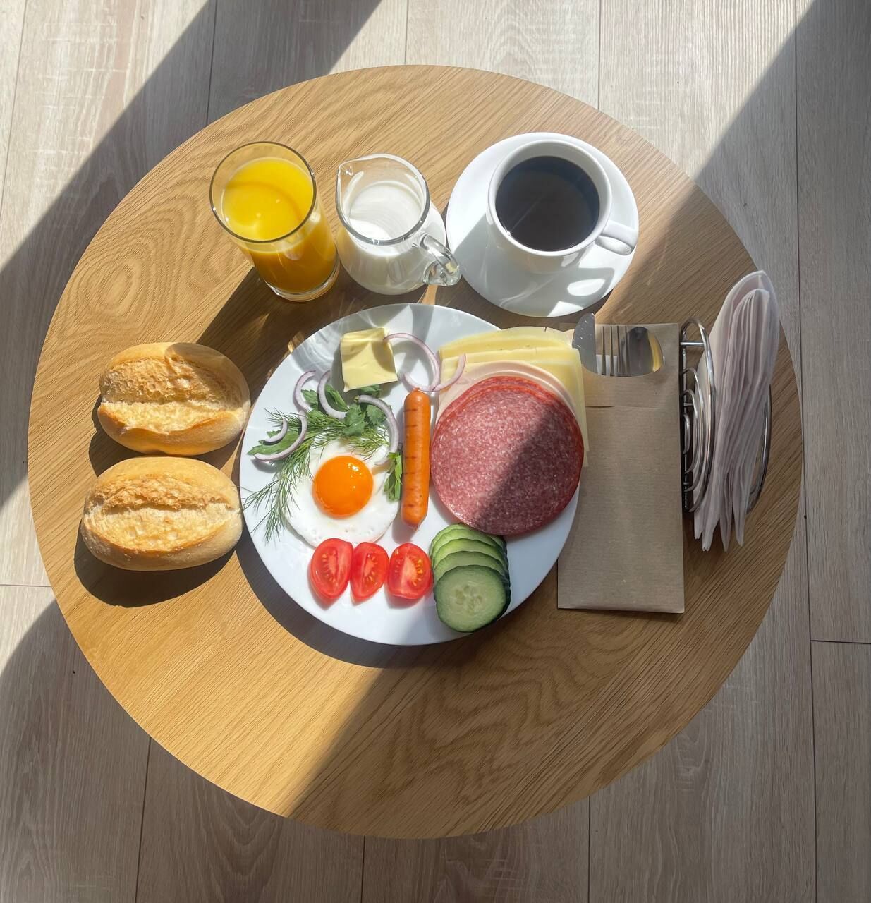 Bandeja de desayuno con huevos, salchicha, queso, tomates, pepinos, pan y bebidas sobre una mesa de madera.