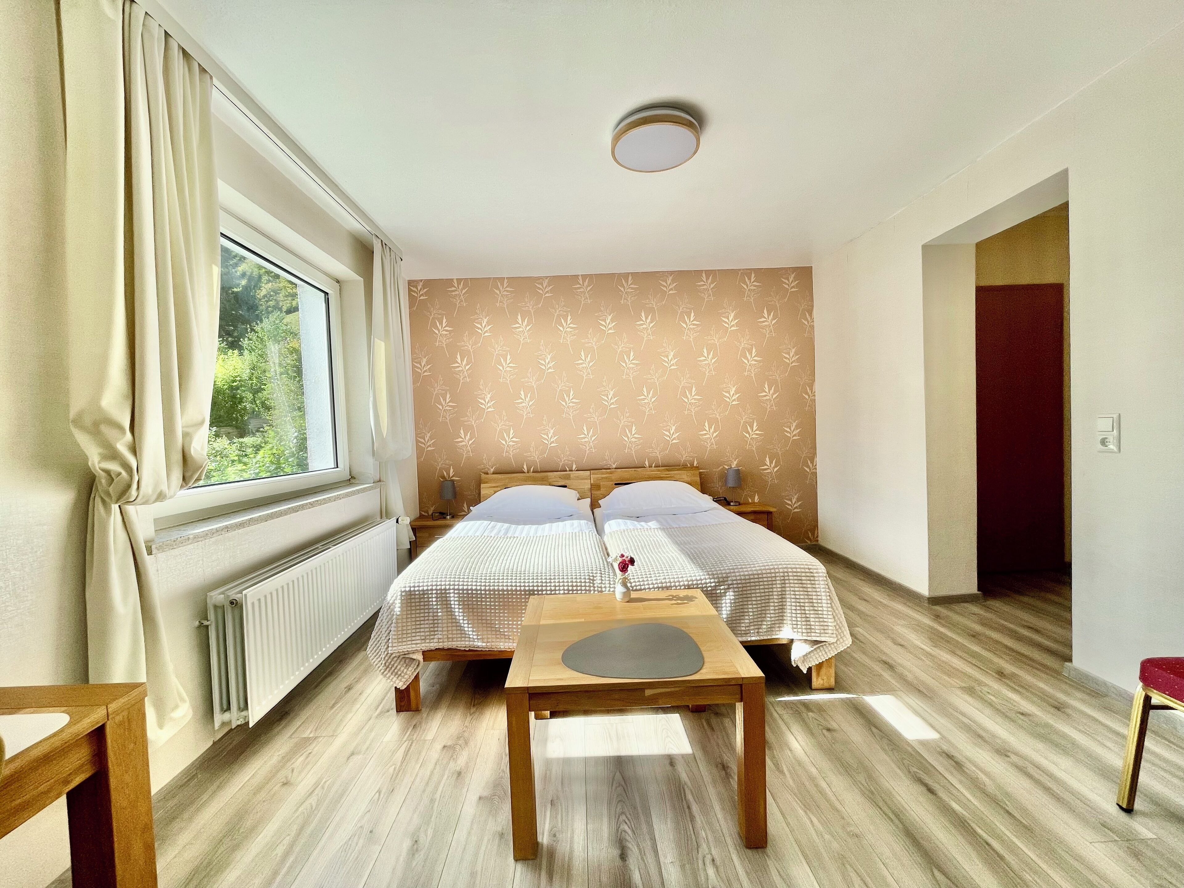 A room with two single beds, a wooden table, a radiator, and a window with curtains.