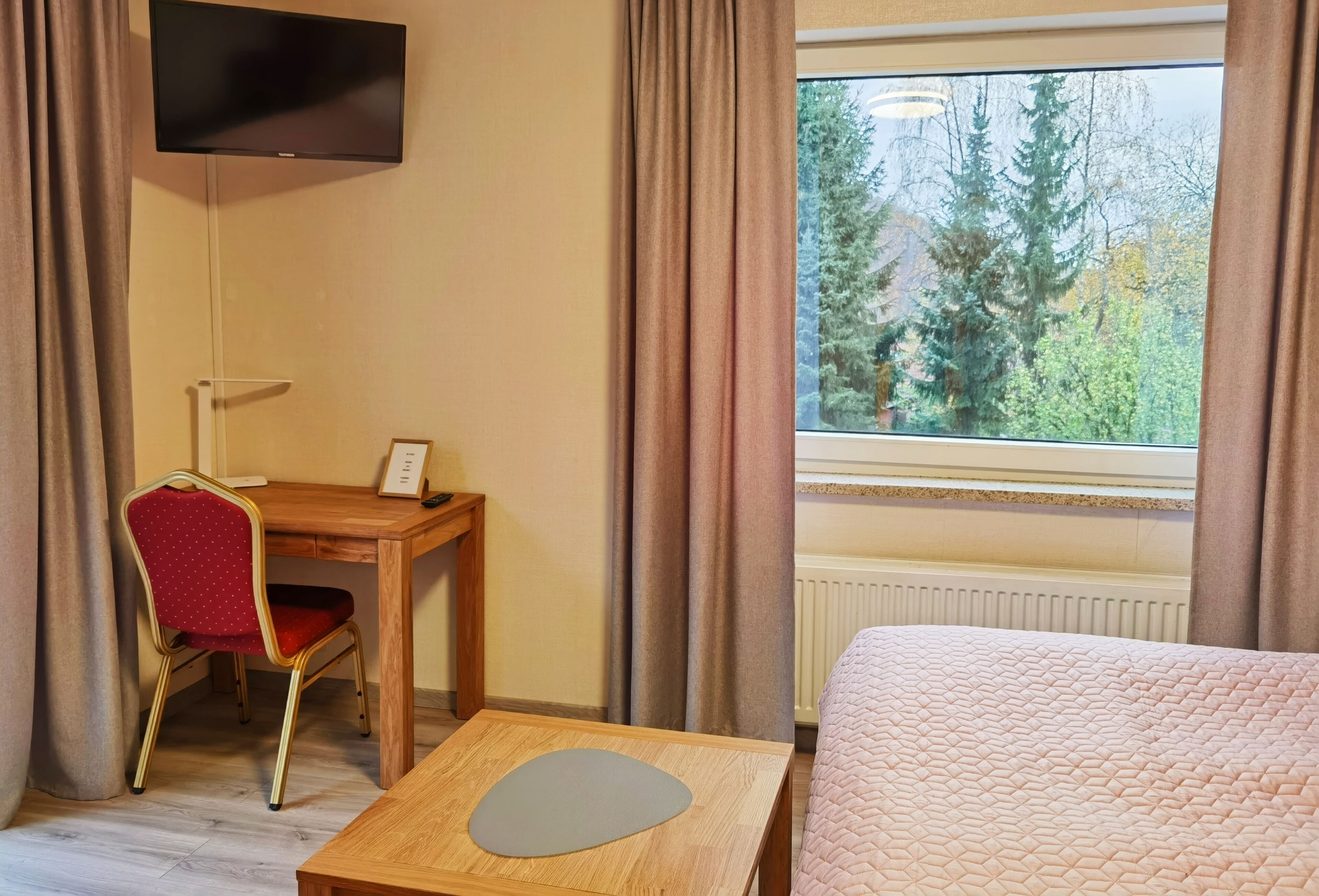 Ein Hotelzimmer mit einem Bett, einem Schreibtisch mit Stuhl, einem an der Wand befestigten Fernseher und einem Fenster mit Blick auf Bäume.