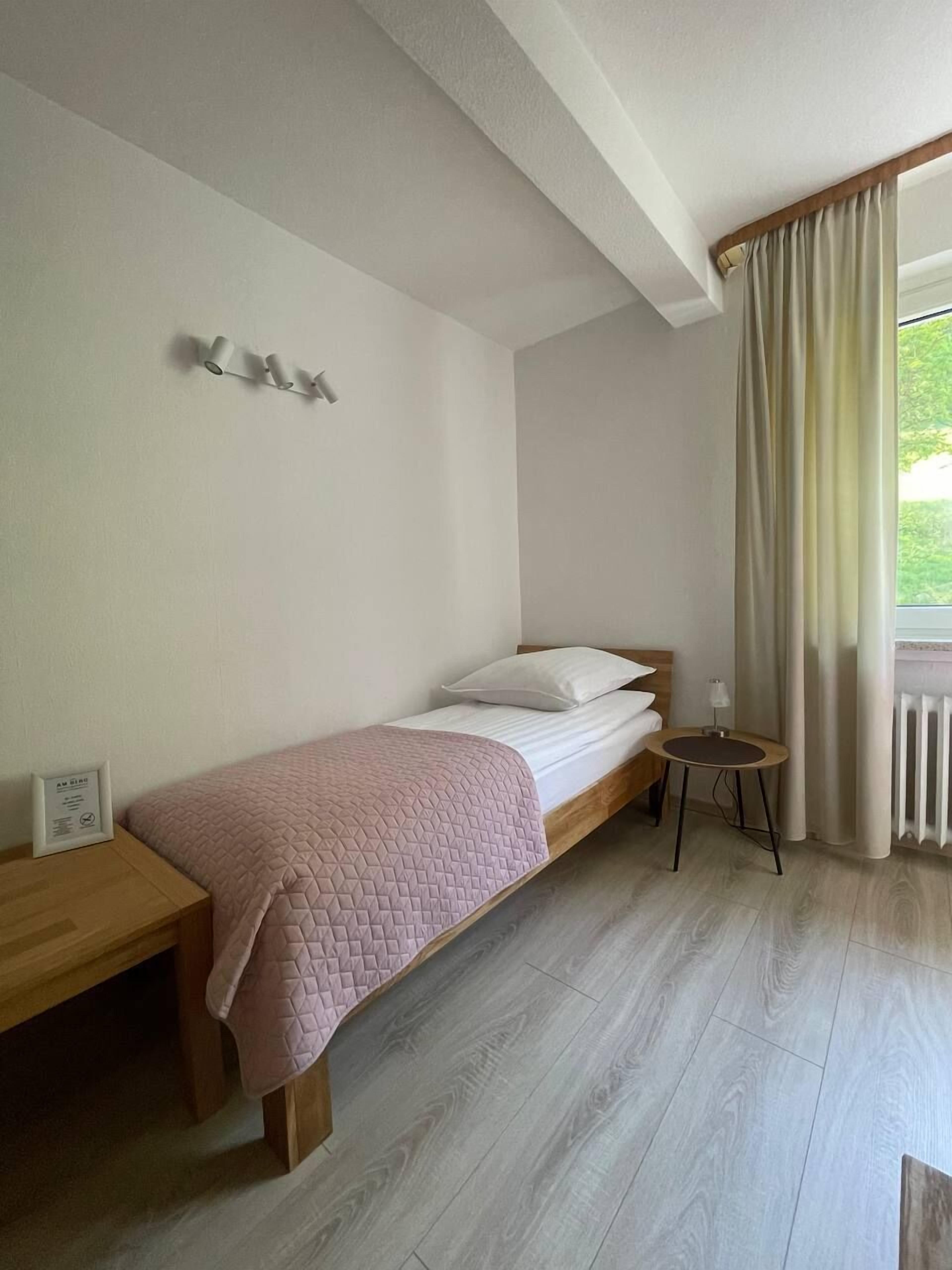 Ein Einzelbett mit einer rosa Steude, ein Nachttisch, ein kleiner runder Tisch mit einer Lampe und ein Fenster mit Vorhängen.