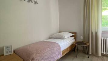 Ein Einzelbett mit einer rosa Steude, ein Nachttisch, ein kleiner runder Tisch mit einer Lampe und ein Fenster mit Vorhängen.