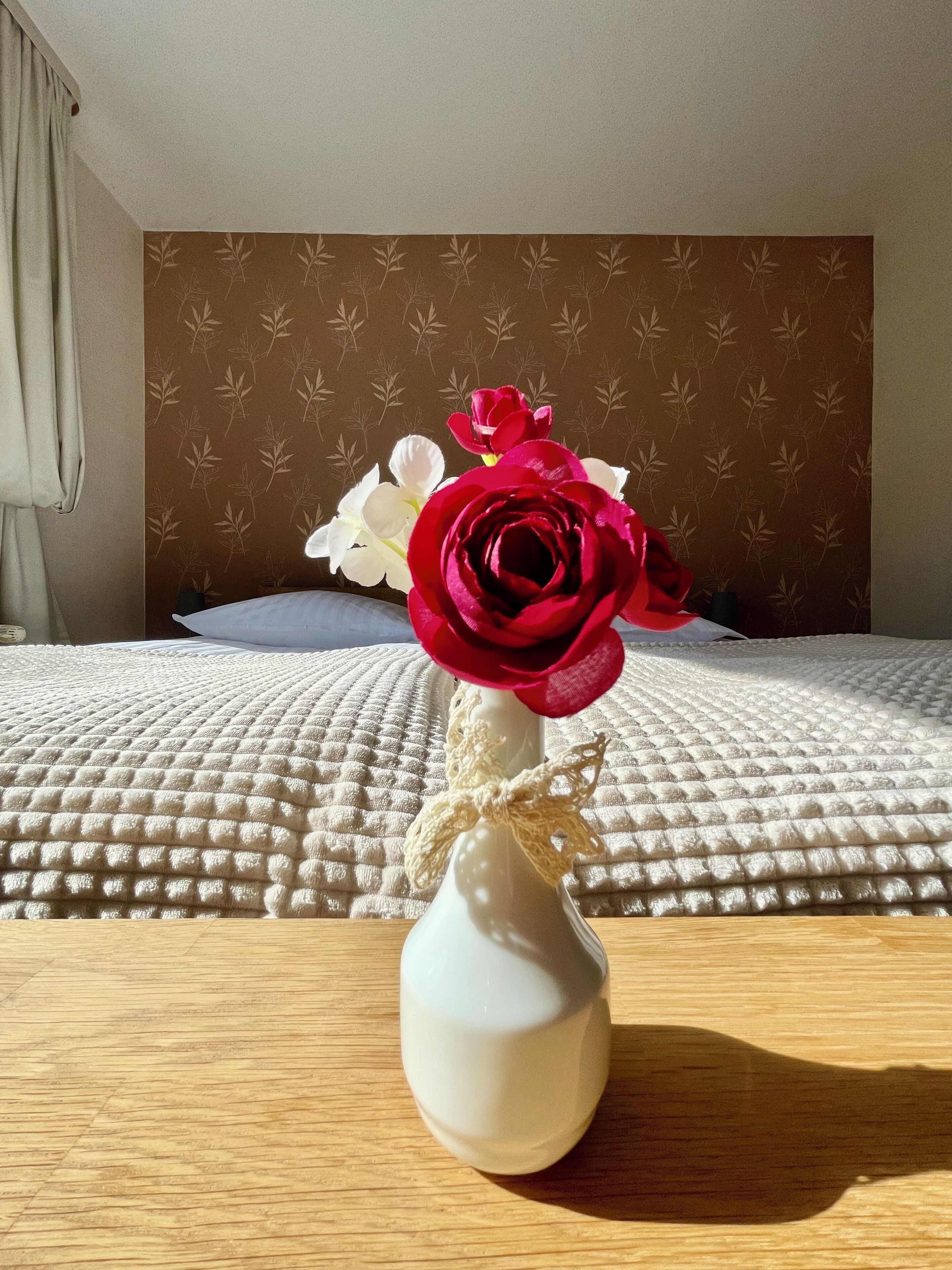En hvid vase med en rød rose og hvide blomster på et træbord i et rum med en seng og tapet.