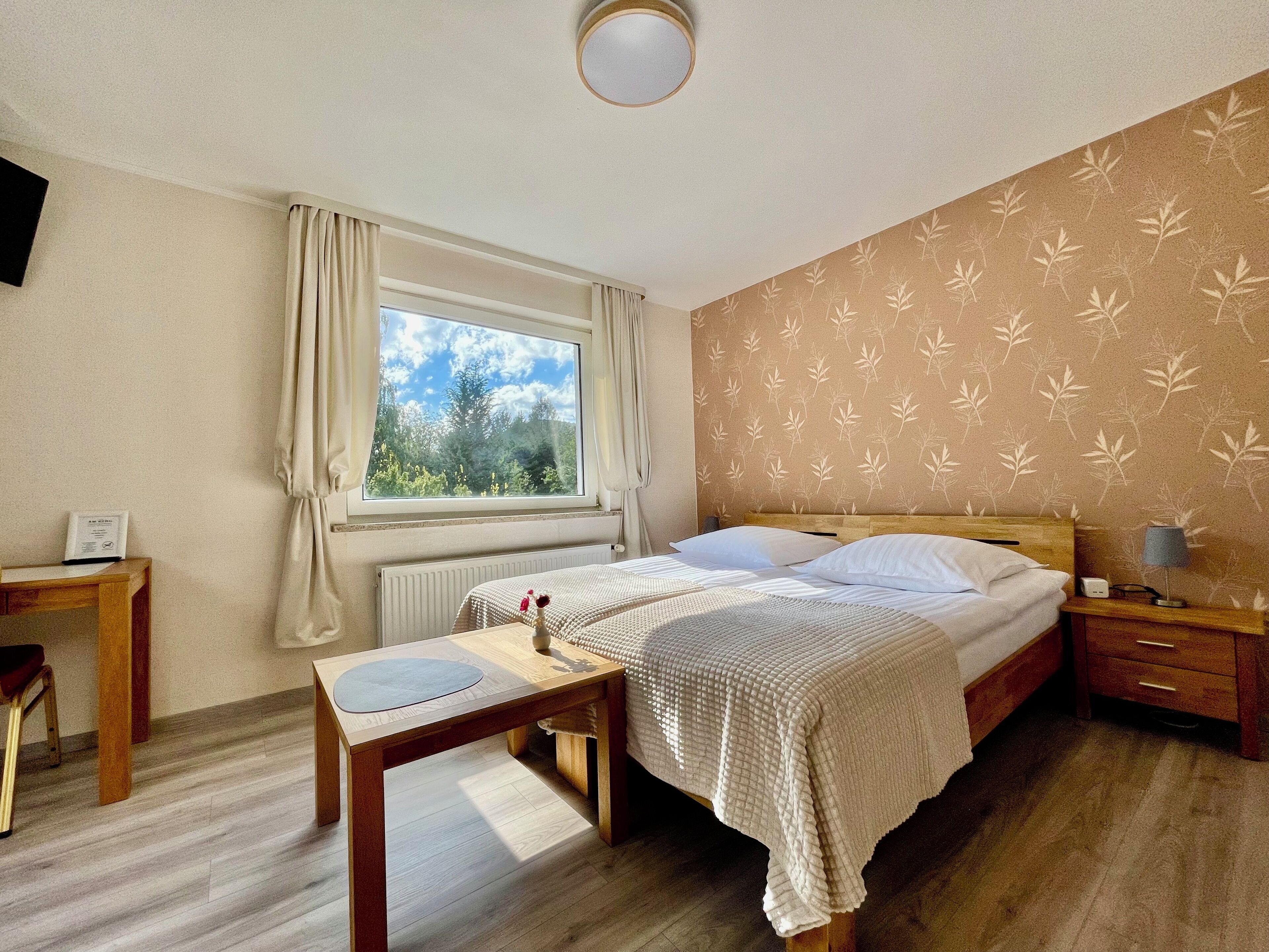 Ein Schlafzimmer mit Bett, Nachttisch, Lampe und Blick ins Freie durch ein Fenster.