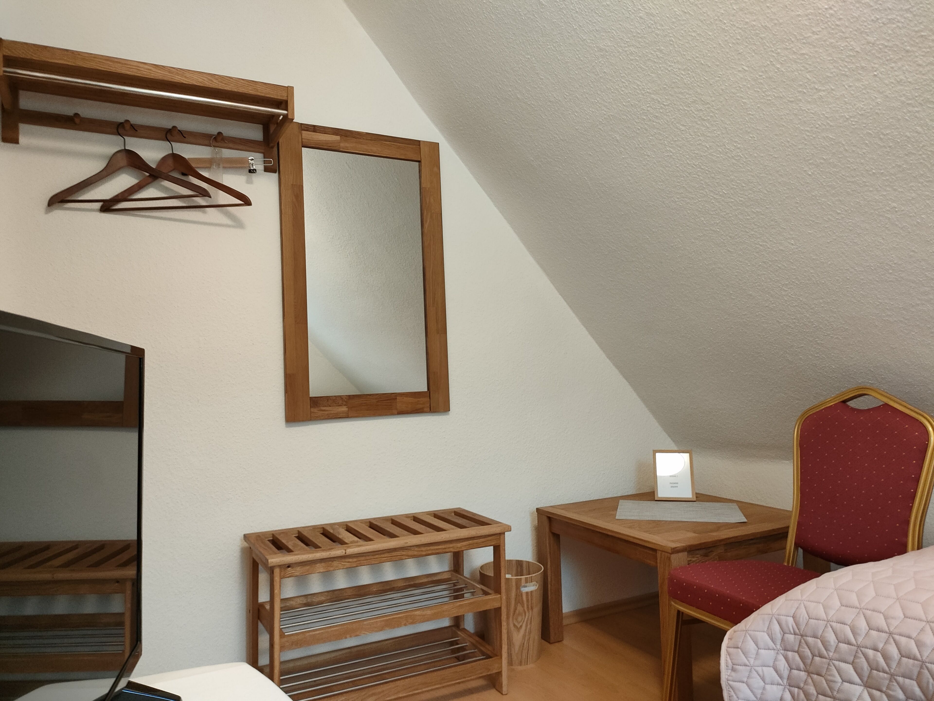 Una habitación pequeña y acogedora con techo inclinado, un perchero de madera con ganchos, un espejo, un estante de madera y un escritorio con silla, además de una cama con cubrecama acolchado.