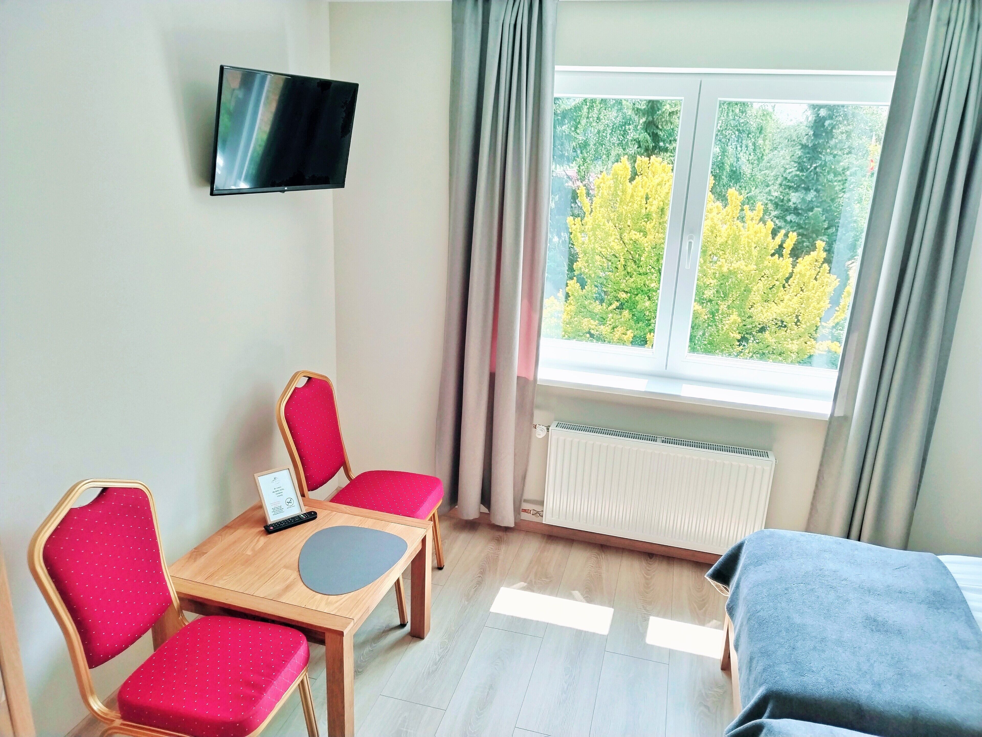 Ett hotellrum med en säng, en tv som är monterad på väggen, ett bord med stolar och ett fönster med gardiner.