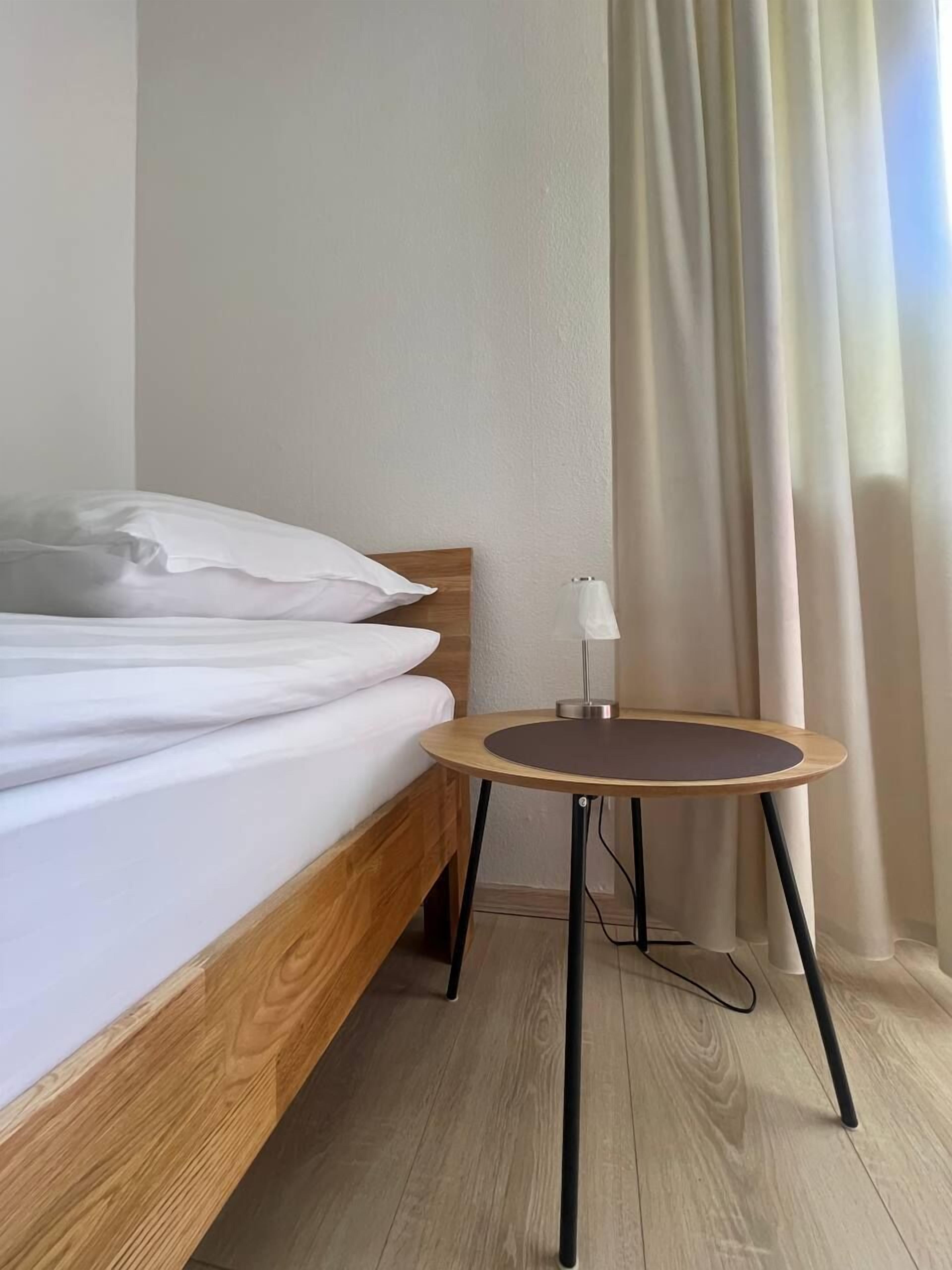 A neatly made bed with white linens, a round wooden table with a lamp, and a window with curtains.