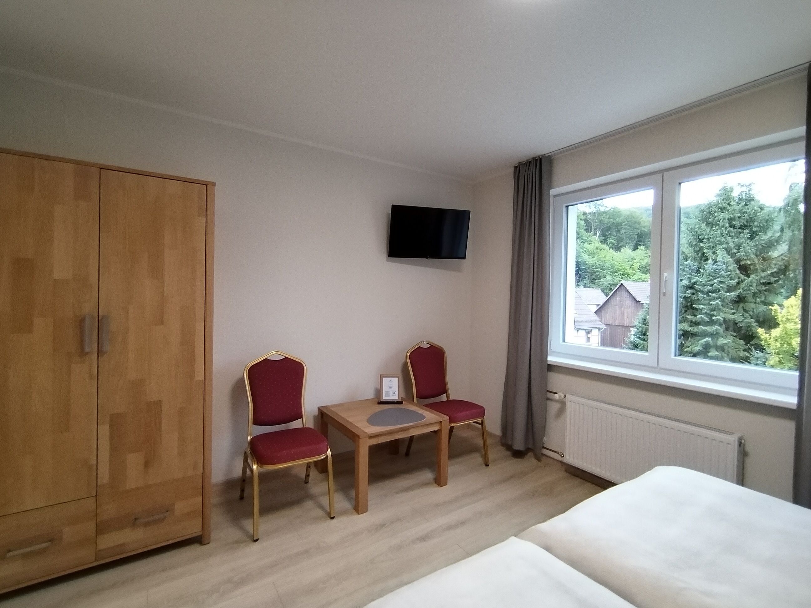 A hotel room with a wooden wardrobe, a TV mounted on the wall, a small table with two red chairs, and a large window with a view of trees and houses.