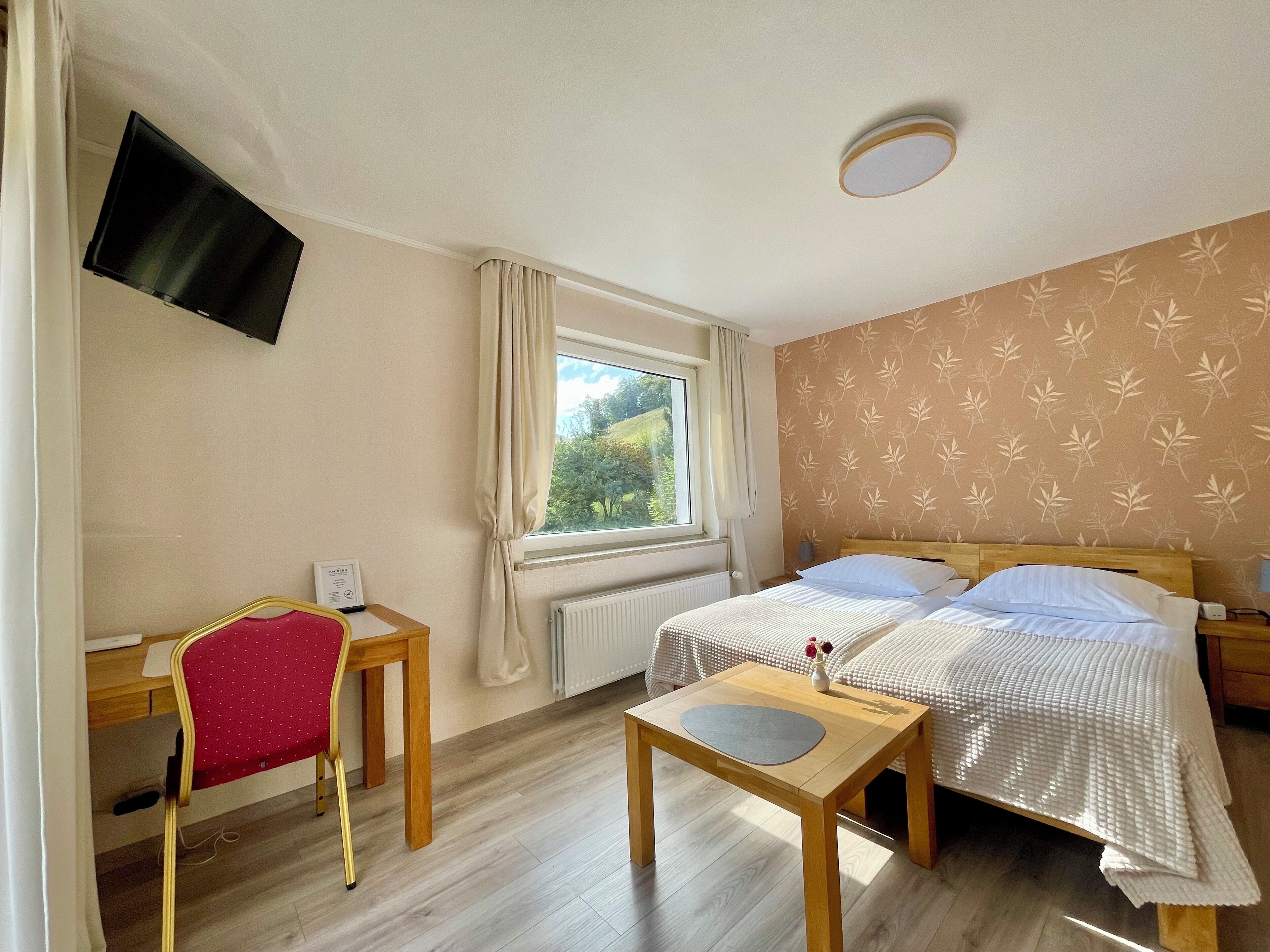 A bedroom with a bed, a desk, a chair, a small table, a TV mounted on the wall, and a window with curtains.
