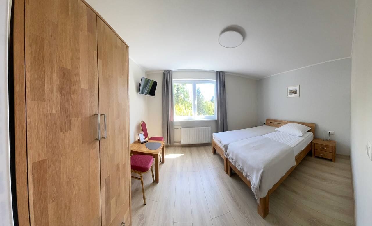 A bedroom with a bed, a desk with a chair, a wardrobe, a window with curtains, and a wall-mounted TV.