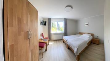 A bedroom with a bed, a desk with a chair, a wardrobe, a window with curtains, and a wall-mounted TV.