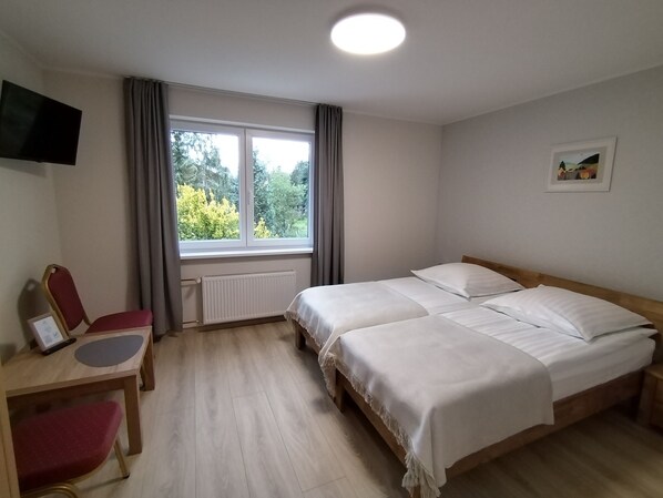 Habitación de hotel con dos camas, un escritorio con silla, un televisor montado en la pared y una ventana con vistas a árboles.
