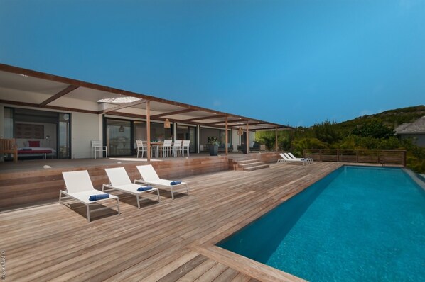 Un espace piscine extérieur moderne, doté d’une terrasse en bois, de chaises longues et d’un coin salon abrité.