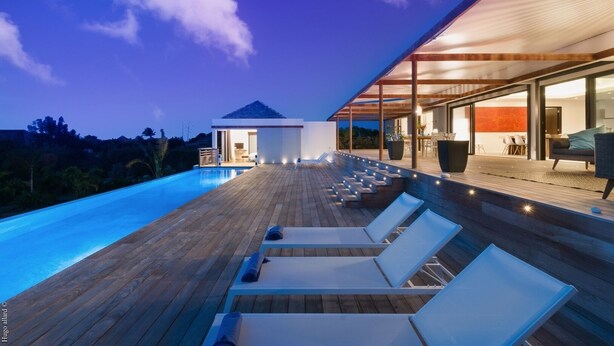 A modern outdoor pool area with wooden decking and lounge chairs, a covered seating area, and a building with large windows. - Cairn (3/4 Rooms) Luxury villa (Petit Cul de Sac)