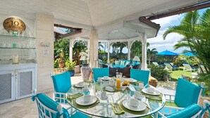 Ein Esstisch mit Glasplatte und blauen Stühlen, umgeben von einer überdachten Terrasse mit Blick auf den Garten und Palmen.