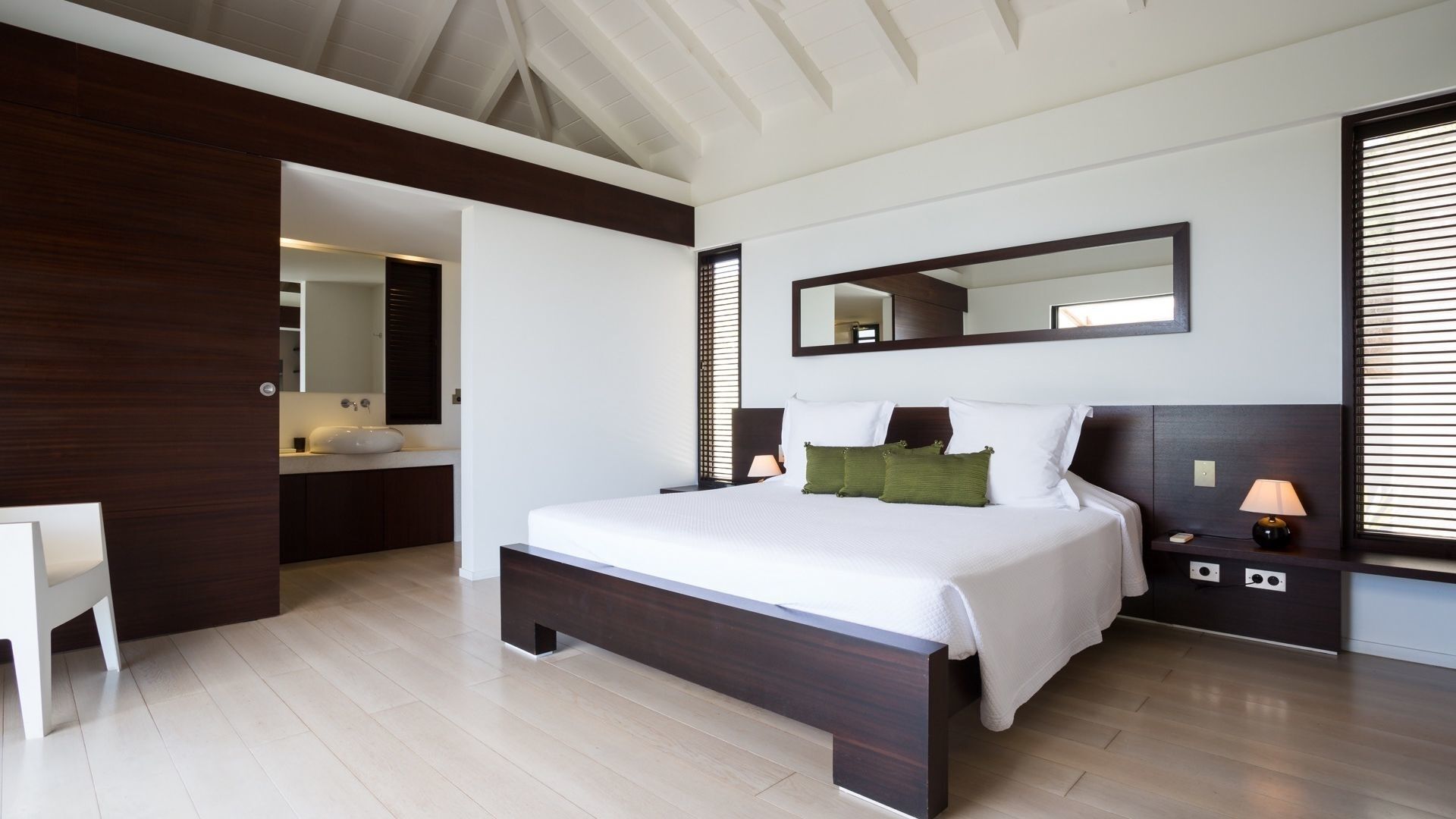 A modern bedroom with a large bed, a bedside table with a lamp, a mirror above the bed, and a view of the bathroom.