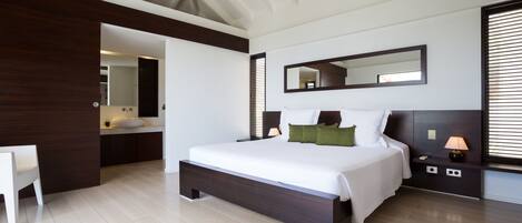 A modern bedroom with a large bed, a bedside table with a lamp, a mirror above the bed, and a view of the bathroom.