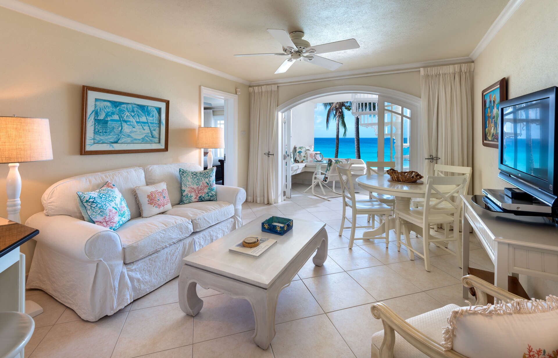 A spacious living room with a white sofa, a coffee table, and a television. There is a balcony with a view of the ocean.
