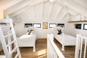 Quarto com duas camas brancas, piso de madeira e teto inclinado com vigas aparentes.
