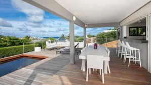 A covered outdoor area with a wooden deck, a pool, and a dining setup with white chairs and a table.