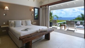 Quarto com uma cama grande, um banco de madeira e vista para o oceano através de uma porta de vidro deslizante.