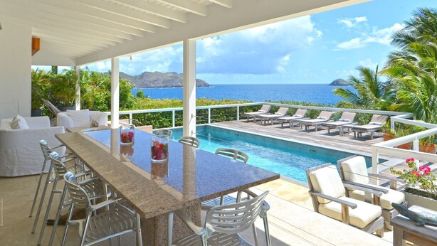 Uma área à beira da piscina com vista para o oceano, um bar com assentos e um terraço coberto.