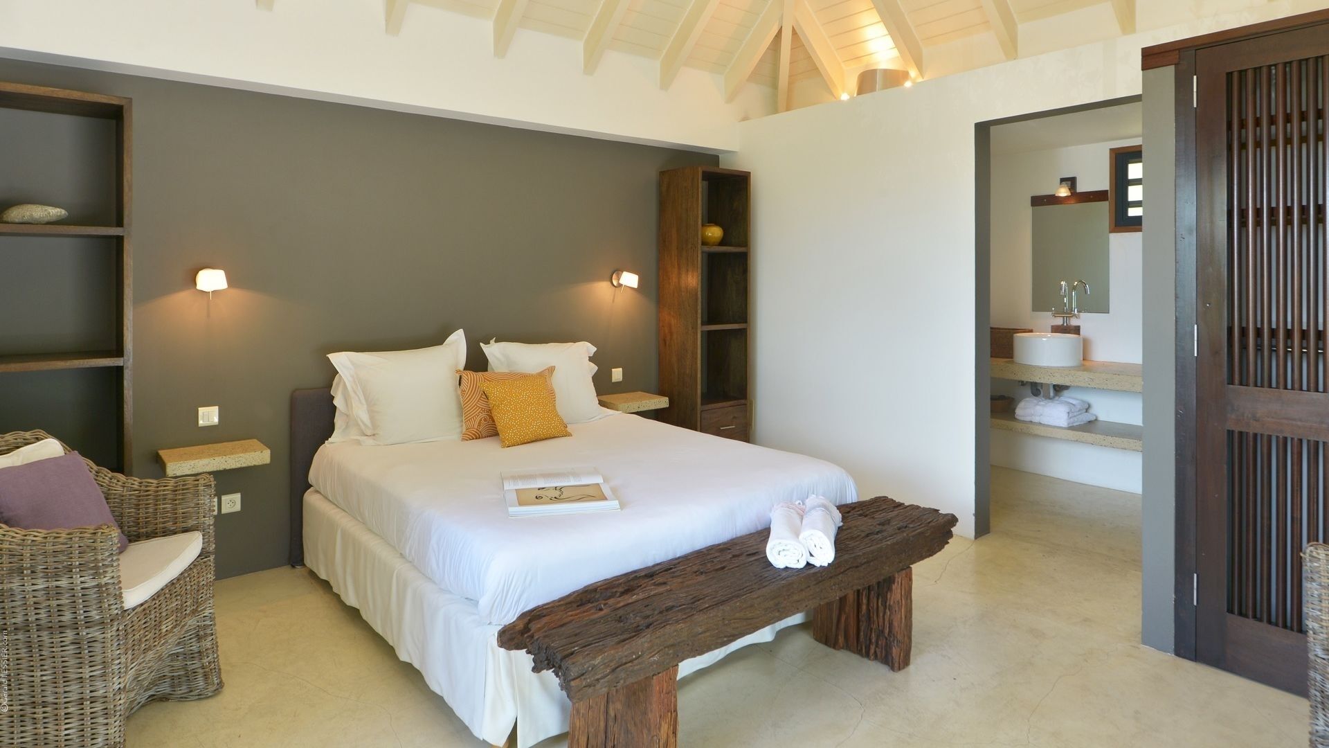 Une chambre avec un grand lit, des tables de chevet, un fauteuil en osier et un banc en bois.