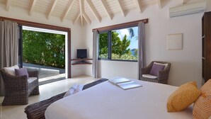 Ein Schlafzimmer mit einem großen Bett, zwei Korbstühlen, einem Fernseher und einem Balkon mit Blick auf Grünflächen und das Meer.