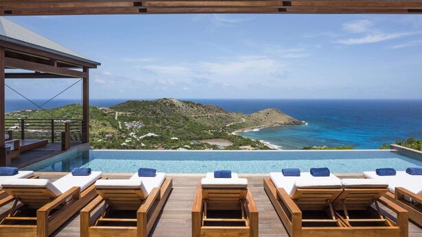 Ein Infinity-Pool mit Holzliegen und Blick auf das Meer und die Hügel.