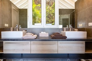 Une salle de bain moderne avec deux lavabos, un grand miroir et une fenêtre donnant sur un espace verdoyant.