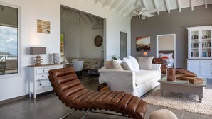 Een moderne woonkamer met een bruine leren fauteuil, een witte bank en een glazen salontafel. Er is een witte kast met boeken en een schilderij van een stadsgezicht.