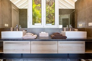 A modern bathroom with two sinks, a large mirror, and a window with a view of greenery.