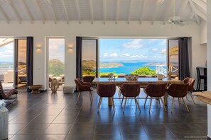 A spacious dining area with a large table and chairs, large windows with a view of the sea, and a ceiling fan.