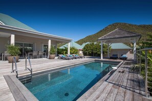 A swimming pool with a wooden deck, lounge chairs, and a covered seating area.