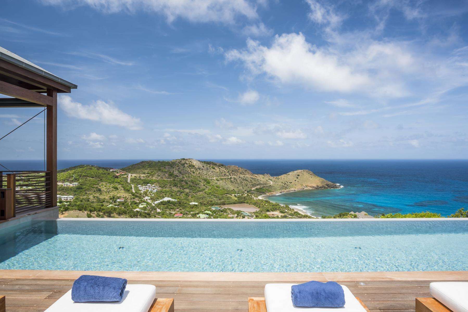 A poolside area with two lounge chairs and a wooden deck, overlooking a coastal landscape with a view of the ocean and a mountainous area in the distance.