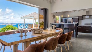 A modern kitchen with dark cabinetry and a central island, a dining area with a wooden table and brown chairs, and a view of the ocean through large windows. - Beautiful Azetes Luxury villa (Vitet)