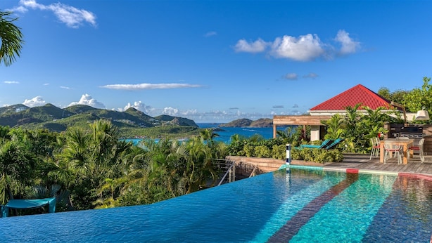 Una piscina con vistas a montañas y al mar, un edificio de techos rojos y exuberante vegetación.