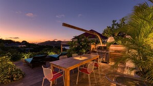 Un área de comedor al aire libre con mesa y sillas de madera, una parrilla y vistas del atardecer sobre un paisaje montañoso.