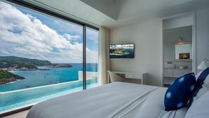 A hotel room with a large window overlooking a coastal view, a bed with white linens and blue pillows, a flat-screen TV on the wall, and a minimalist bathroom with a mirror and sink.