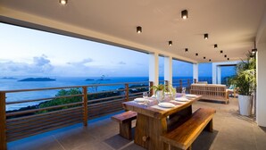A spacious balcony with a wooden table set for dining, a bench, and a seating area with a view of the ocean and islands.
